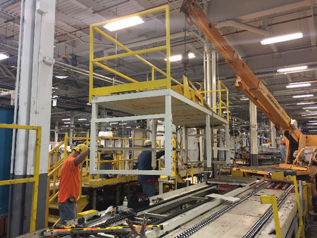 men using a crane to lower a metal structure on to a conveyor in a factory